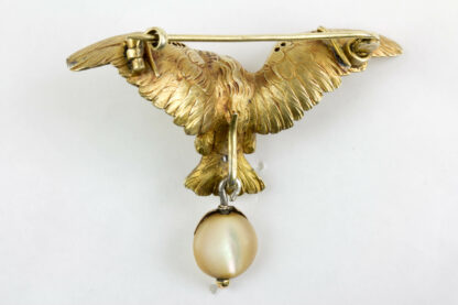 Brosche, um 1900, in Form eines Adlers, Silber vergoldet, Augen mit kleinen Rubinen besetzt, Flügelkanten mit Rosendiamanten, anhängend eine Perlmutscheibe in Rotgold gefasst, sehr feine Ausführung. H: 4 cm, B: 5,7 cm.